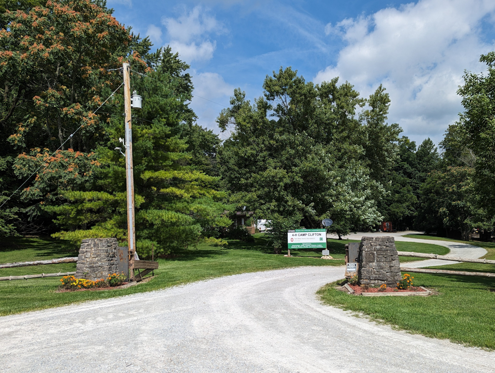 A paved entrance road flanked by stone pillars, with a large sign reading "4-H Camp Clifton"