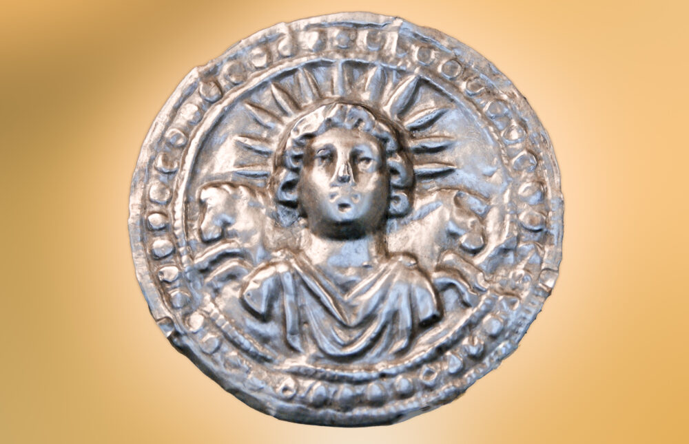 Against a blurred background, a silver disc depicting the bust of a young man crowned with the rays of the sun