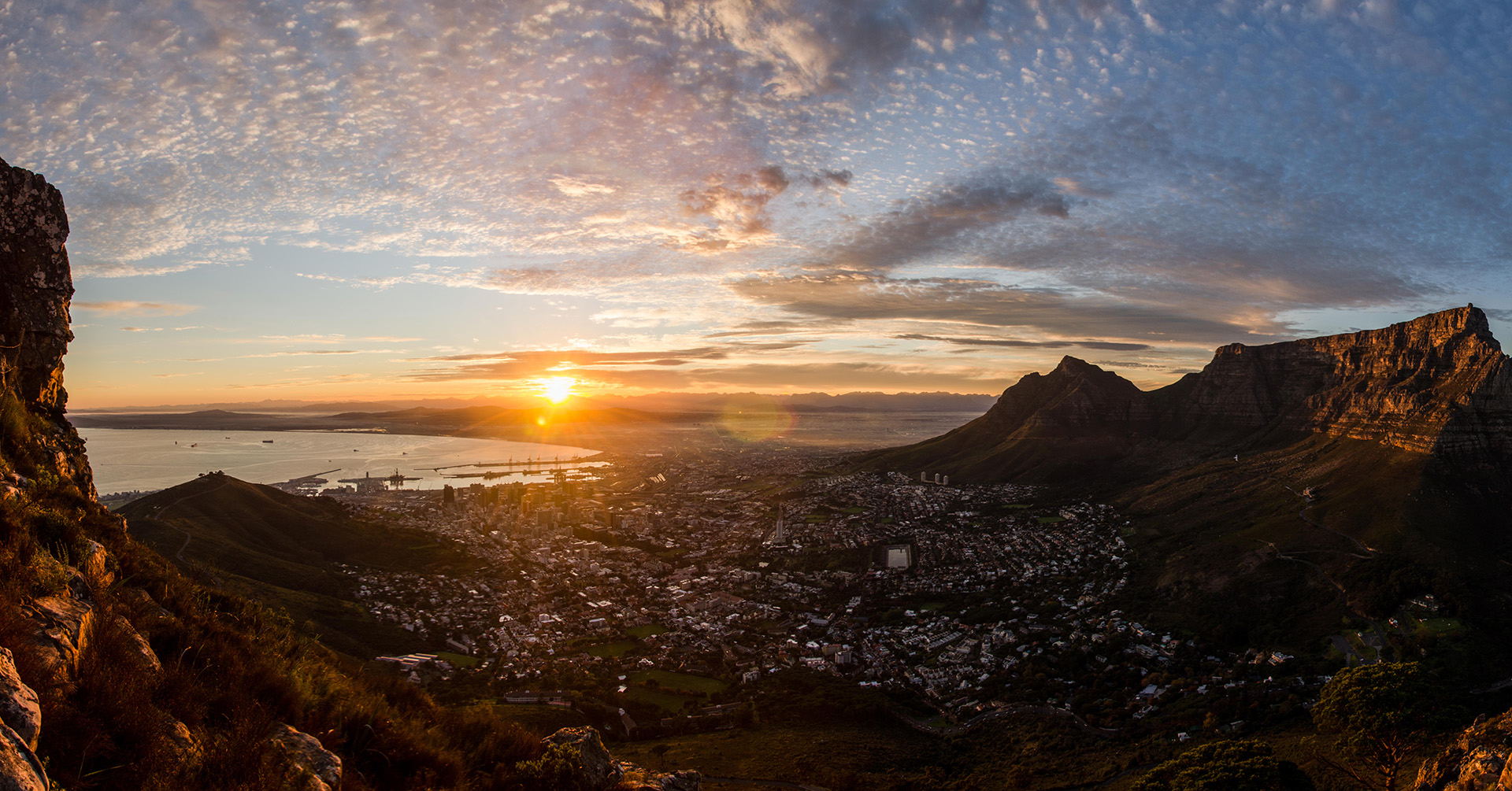 The brilliant sun rises through a skirt of clouds, illuminating a still ocean bay and a town nestled among mountainous outcrops.